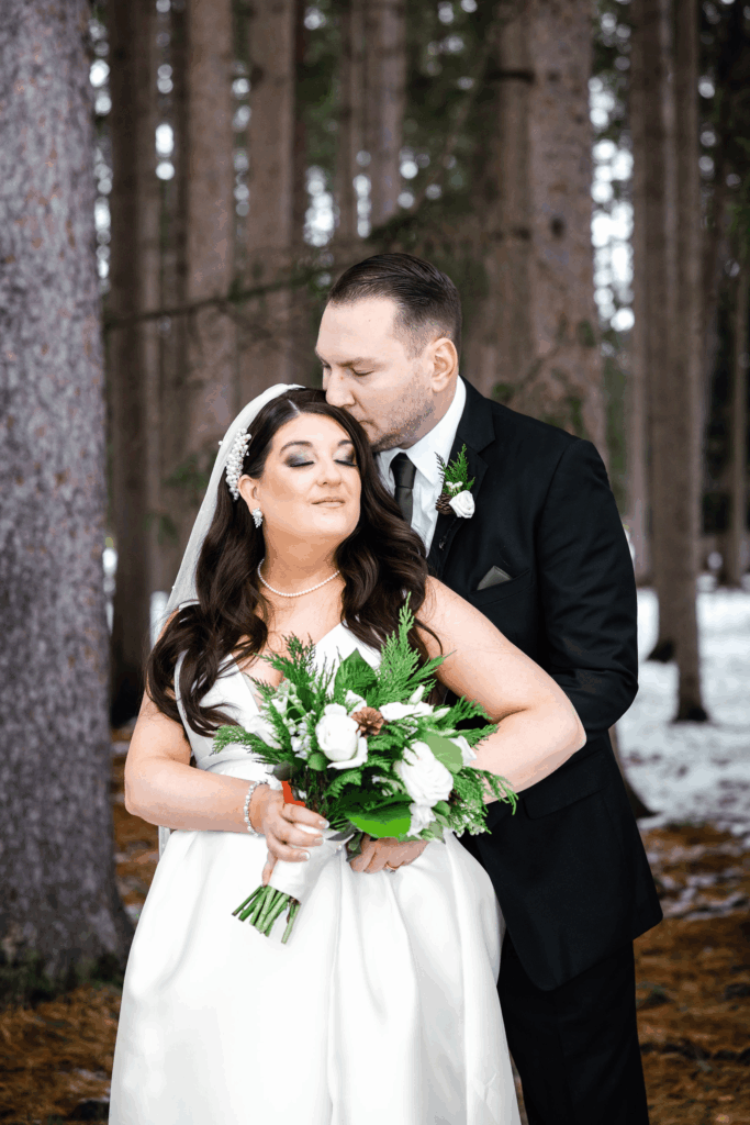 Bride and groom posing for their wedding photos. Groom is kissing the brides head they are surrounded by snow and trees.